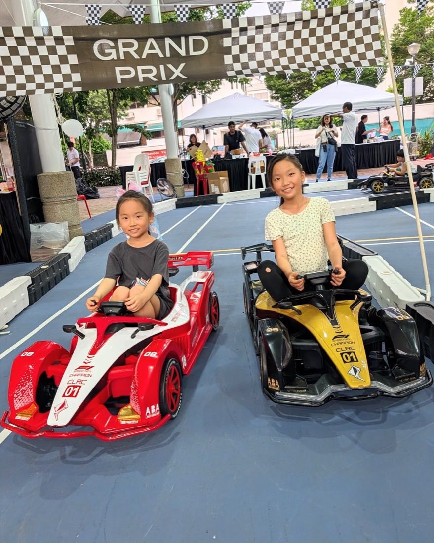 Two young drivers on the grid in red and gold ride-on race cars under the Grand Prix start banner